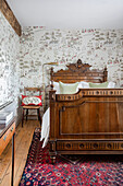 Wooden bed with carvings and patterned wallpaper in bedroom with carpet and wooden floorboards