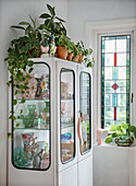 Display cabinet with plants, vintage crockery and vases next to stained glass window
