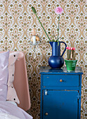 Bedside table in blue with vase, flowers and cactus in front of retro wallpaper