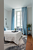 Schlafzimmer mit weißer Bettwäsche, hellblauen Vorhängen und Parkettboden