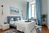Blaues Bett mit passenden Vorhängen und Meeresbild im hellen Schlafzimmer