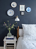 Bedside table with glass vase and flowers next to bed in front of dark blue wall with wall plates and picture frames