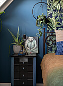 Bedside table with plant, lamp and artwork in front of blue-patterned wall