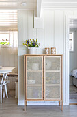 Sideboard aus Holzgeflecht mit Blumenvase und Teelichtern vor weißer Vertäfelung im Flur