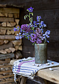 Bouquet of meadow flowers in a metal vase on a rustic wooden table in front of a stack of wood