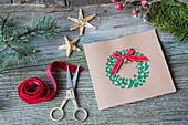 Homemade Christmas card with wreath motif, surrounded by handicraft utensils and fir branches