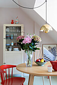 Round dining table with bouquet of flowers, red chair and wooden display cabinet in light-coloured room