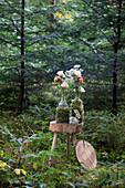 Mit Moos umhüllte Glasflaschen mit Rosen und Schleierkraut auf rustikalem Holztisch im Wald