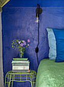 Bedside table with books and bouquet of flowers, bed with green blanket, blue wall