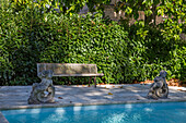 Stone bench and gargoyle statues by the pool in front of an evergreen hedge