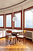 Round dining table with wooden chairs and arc lamp in front of many windows