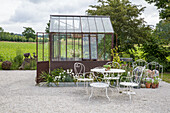 Romantic seating area with white iron furniture in front of greenhouse in the garden