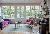 Living room with pink armchair and couch and wood-burning stove in front of large windows overlooking the garden