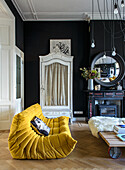 Yellow designer sofa and black walls in living room with antique cupboard and fireplace