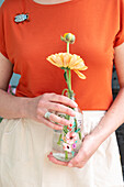 Frau mit orangefarbenem T-Shirt hält Gerbera in Glasvase mit Blumenmuster