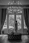 Frau in langem Kleid blickt aus dem Fenster eines noblen Zimmers mit Kronleuchter