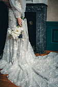 Braut in besticktem weißen Hochzeitskleid mit Brautstrauß vor Kamin