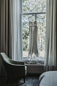 Wedding dress hangs on window in room with green armchair and curtains