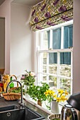 Küchenfenster mit Raffrollo und frischen Blumen und Kräutern auf der Fensterbank