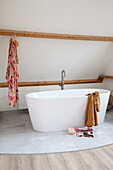 Freestanding bathtub, wooden strips and magazine on grey tiles in bathroom with sloping roof