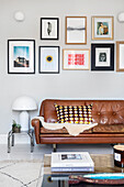 Picture wall above brown leather sofa with patterned cushions and stack of books on glass table