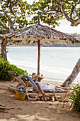 Woman enjoying an idyllic holiday on the beach on a sun lounger under a thatched umbrella