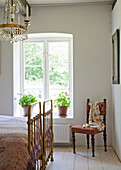 Bright bedroom with brass bed, antique chair, crystal chandelier and potted plants by the window