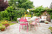 Tisch und bunte Gartenstühle auf sonniger Terrasse umgeben von Pflanzen