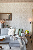 Grey corner sofa with cushions and blanket in front of patterned wallpaper, cat lying on it