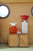 Two ornate table lamps on a wooden chest of drawers in front of porthole windows