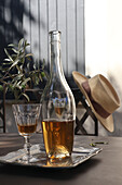 Bottle and glass with drink on table, olive branch, chair with straw hat in background