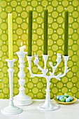 White candle holders with green candles in front of green patterned wallpaper