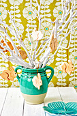 White branches with wooden bunny pendants in a green vase in front of patterned wallpaper