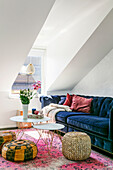 Living room with blue velvet sofa, poufs and colourful carpet under a sloping roof