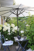 Small garden alcove with bamboo umbrella and hydrangeas, anthracite-coloured folding furniture on gravel path