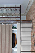 Metal loft bed with wrought-iron railing above wardrobe with open curtain