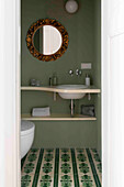 Bathroom with patterned tiled floor and round mirror with gold frame on green wall