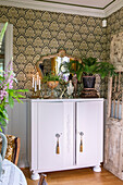 White sideboard in front of baroque patterned wallpaper with plants and candlestick