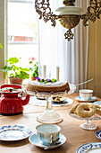 Breakfast table with glass pedestal dish, pastries and red tea kettle in front of window with curtain