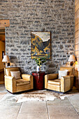 Two leather armchairs and round side table in front of stone wall with painting