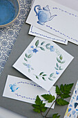 Hand-painted watercolour cards with blue flower and pitcher motifs on a grey background