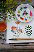 Watercolour drawing with sparrow on branch and marigold flowers, surrounded by painting utensils