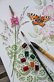Watercolour painting of butterfly on dog rose with painting utensils