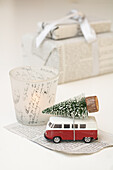 Miniature van with a Christmas tree next to a candle and presents in newspaper