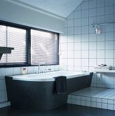 Dark louver blinds above black-clad bathtub and white-tiled platform in austere bathroom