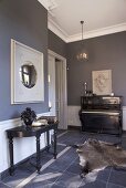 Grey-blue interior with black console table, black antique piano, animal-skin rug, grey-blue floor tiles and round convex mirror