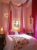 Doppelbett unter beleuchtete Lampe mit religöser Zeichnung an eine rosa Wand im Schlafzimmer mit pinken Stoffen und Vorhängen