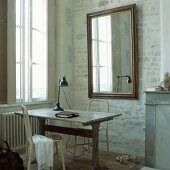 Ein antiker Holztisch und zwei weiße Stühle vor einem Wandspiegel in der Ecke eines Raums mit Backsteinwänden