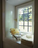 Gestapelte Handtücher mit duftender Seife, daneben elegante Badeutensilien und eine üppige Blüte auf weisser Fensterbank