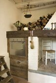 Corner of a kitchen with built-in stainless steel oven in a rustic wooden cabinet next to a chimney and a place to store firewood